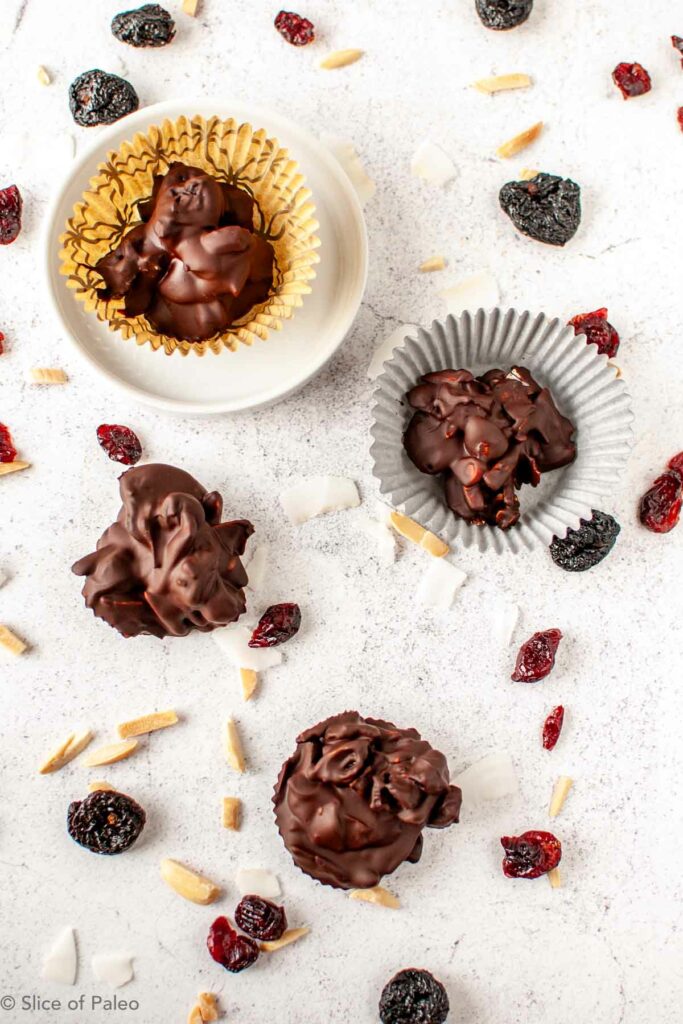 Paleo chocolate clusters displayed on a counter with dried fruits, nuts, and coconut chunks.